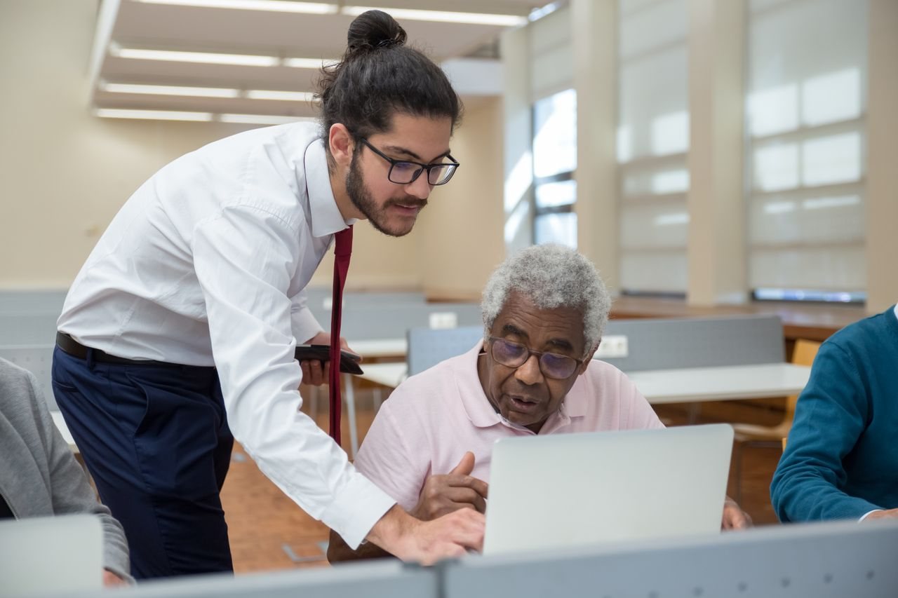 Free A young man helps a senior adult with laptop use, emphasizing tech education. Stock Photo
